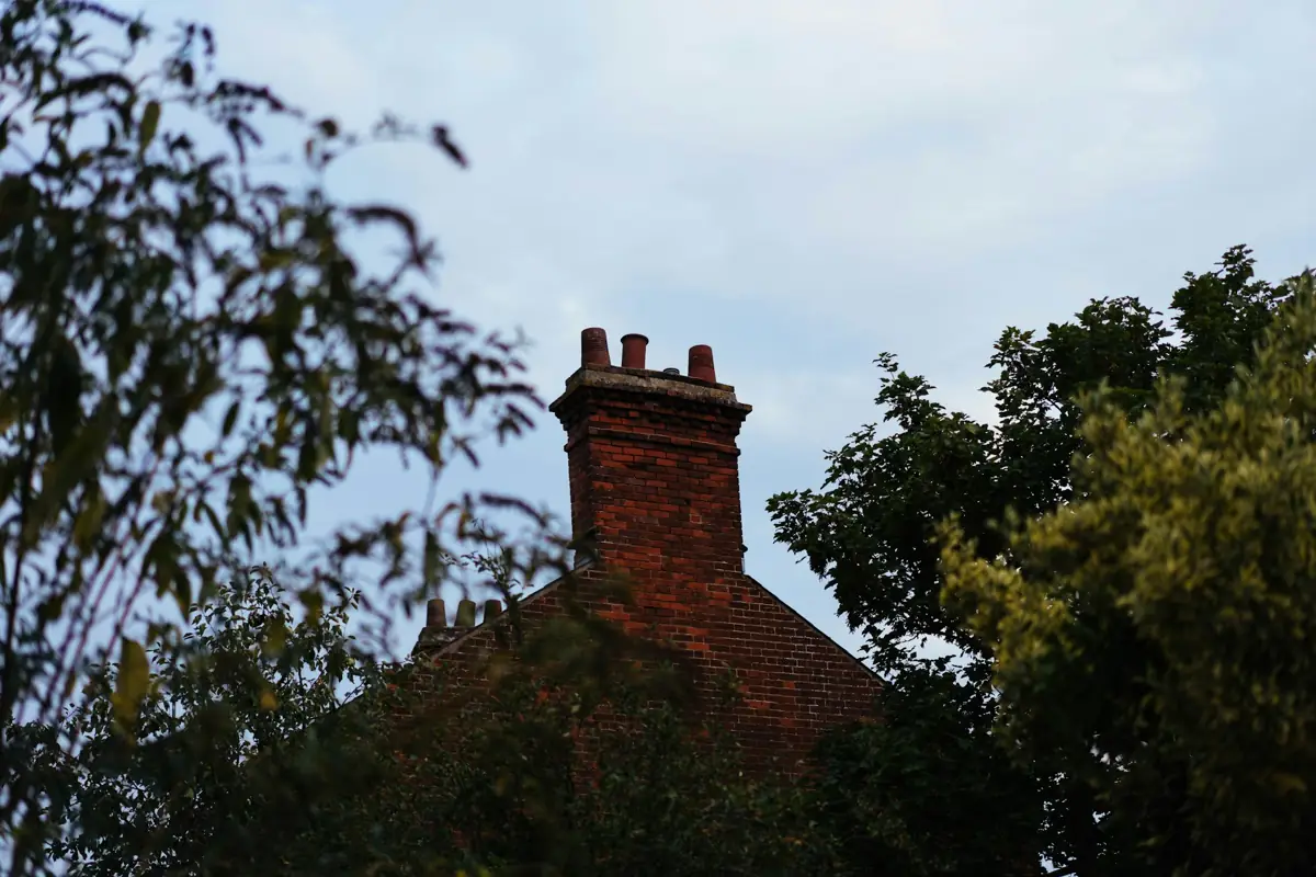 A red brick chimney with clay pots rising above a tiled roof and trees